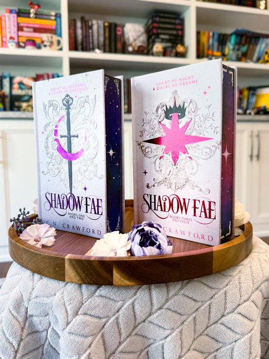 Two fancy special editions novels titled 'Shadow Fae 1-2' and 'Shadow Fae 3-4'  by C.N. Crawford. Books are displayed on a wooden tray with decorative items, against a bookshelf background.